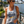 Load image into Gallery viewer, A woman wearing a light gray graphic tank top with 'LET'S GO Wisco' printed on the front, paired with denim shorts, standing outdoors near a waterfront with a city skyline in the background.
