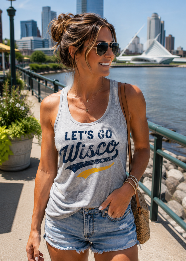 A woman wearing a light gray graphic tank top with 'LET'S GO Wisco' printed on the front, paired with denim shorts, standing outdoors near a waterfront with a city skyline in the background.