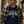 Load image into Gallery viewer, A man wearing a navy blue graphic t-shirt with 'MEET ME AT THE TAVERN WISCONSIN UP' printed on the front, layered under an open brown and beige plaid flannel shirt, and wearing a brown and tan baseball cap. He is standing outdoors in a pine forest next to a rustic log cabin.
