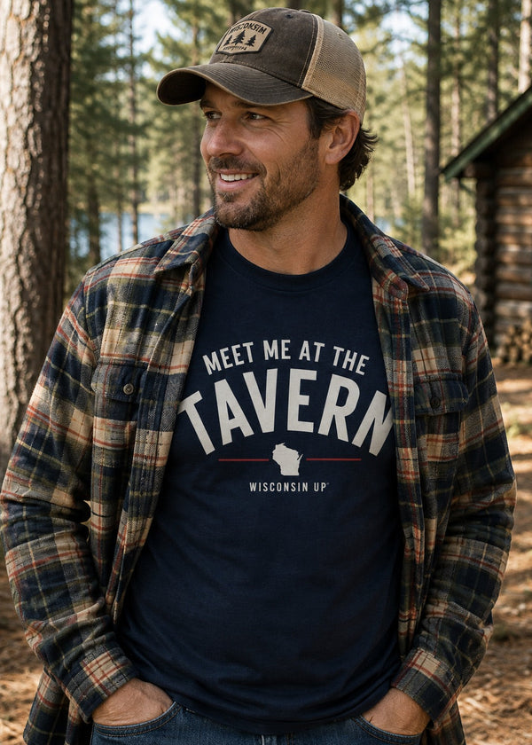 A man wearing a navy blue graphic t-shirt with 'MEET ME AT THE TAVERN WISCONSIN UP' printed on the front, layered under an open brown and beige plaid flannel shirt, and wearing a brown and tan baseball cap. He is standing outdoors in a pine forest next to a rustic log cabin.