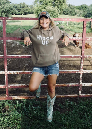 Woman in a hoodie with 'Wesco Up' text, standing next to a metal gate with cows in the background.
