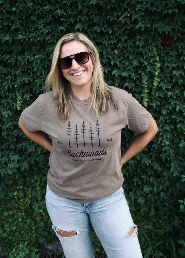Woman wearing a brown t-shirt with a forest design and text, standing against a green ivy wall.