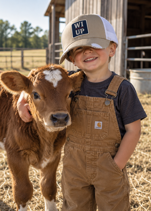 A smiling young boy stands in a barn next to a brown calf. He is wearing a beige and white trucker hat with a 'WI UP' patch, a dark gray t-shirt, and brown Carhartt overalls.