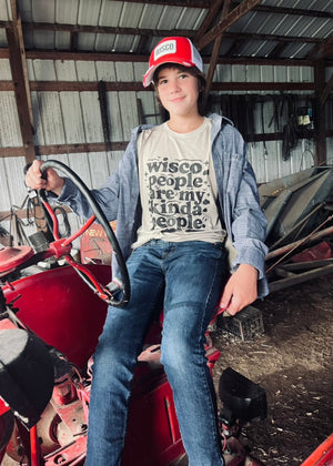 Person sitting on a red tractor wearing a 'Wisco people are my kinda people' shirt in a barn setting.
