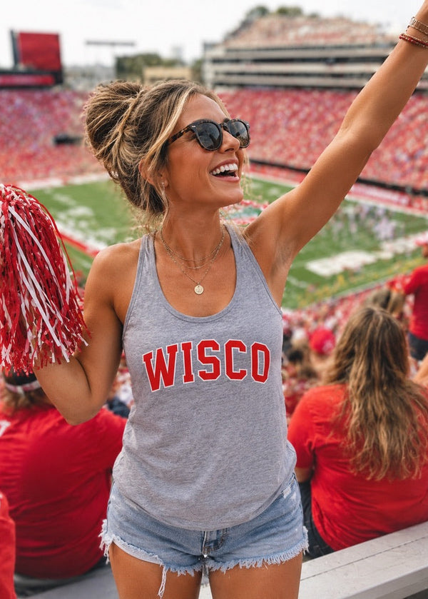 A woman cheering at a stadium wearing a heather gray tank top with 'WISCO' printed in red across the chest, paired with denim shorts and sunglasses. She is holding a red and white pom-pom.