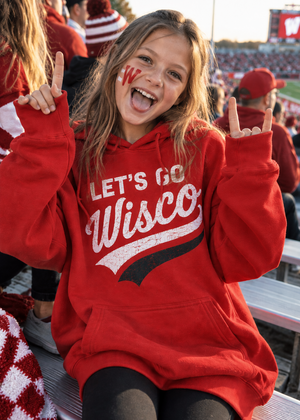 A young girl cheering at a sports event, wearing a red hoodie with 'LET'S GO Wisco' printed in white and black. She has 'W' face paint on her cheek and is sitting in stadium bleachers with other fans in the background.