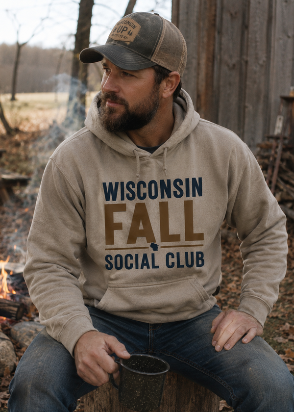 A man wearing a beige hoodie with 'WISCONSIN FALL SOCIAL CLUB' printed on the front, sitting outdoors by a campfire and holding a mug.