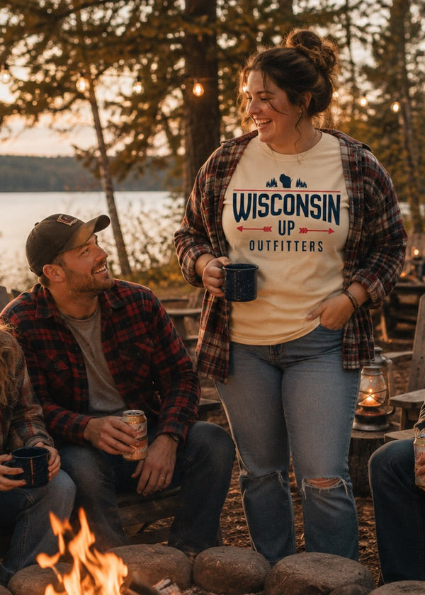 Four friends gathered around a campfire at dusk by a lake, wearing flannel shirts and beanies. The standing woman wears a cream graphic T-shirt that reads 'WISCONSIN UP OUTFITTERS' with a blue mountain-and-river logo.