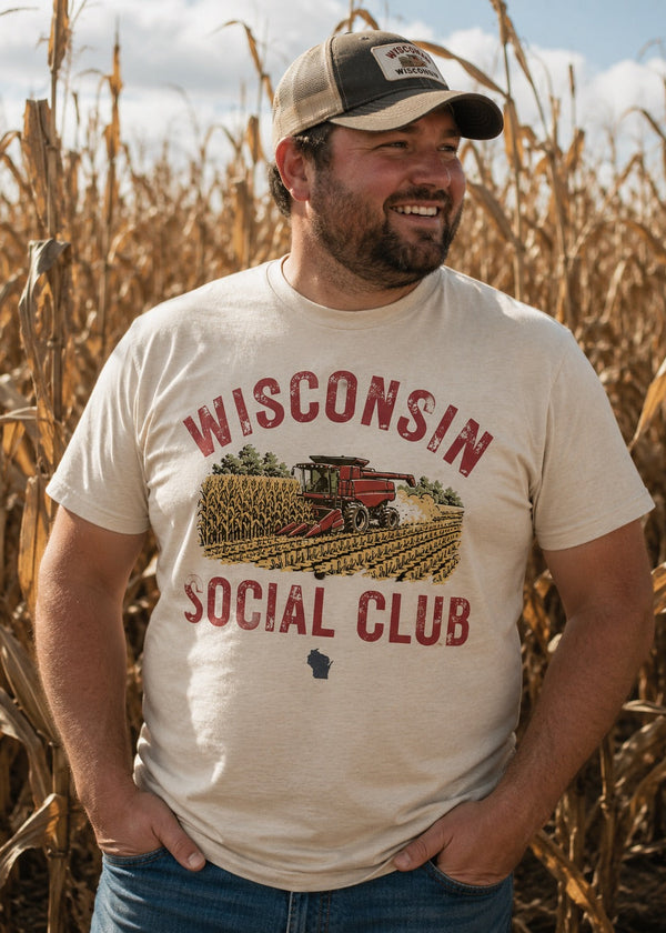 A man wearing a cream-colored graphic t-shirt that reads 'WISCONSIN SOCIAL CLUB' with a combine harvester illustration, standing in a dry cornfield.