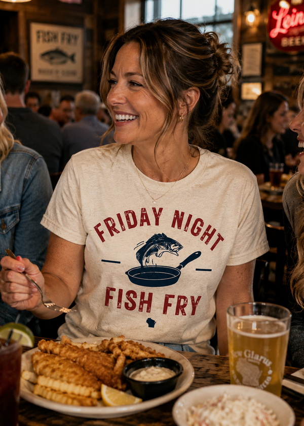A woman smiling at a restaurant table wearing a cream graphic t-shirt that reads 'FRIDAY NIGHT FISH FRY' with a fish frying pan illustration. In front of her is a plate of fish and chips, and a glass of beer on the table.