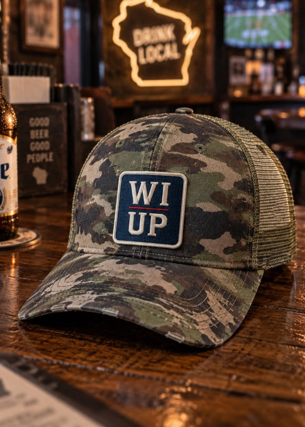 A camouflage trucker hat with a blue and white 'WI UP' patch on the front, displayed on a wooden bar counter next to a Miller Lite bottle in a bar setting.