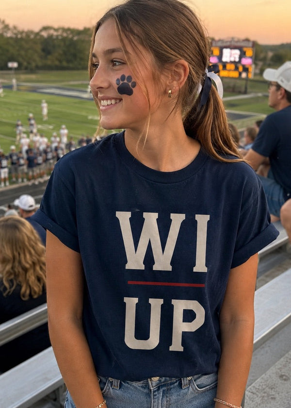 A young woman in a navy blue t-shirt with 'WI UP' printed on the front, sitting in an outdoor stadium at sunset. The shirt features a navy blue background with 'WI' in cream and 'UP' in navy, separated by a thin red line, with a navy paw print graphic on the 'I'.