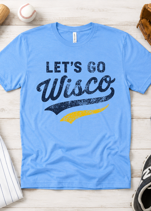 A light blue crewneck t-shirt with a distressed graphic reading 'LET'S GO Wisco' in navy and yellow, laid flat on a light wood background with baseball-themed props.