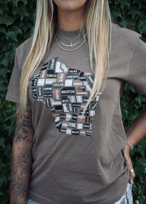 Person wearing a brown t-shirt with graphic design against a green leafy background