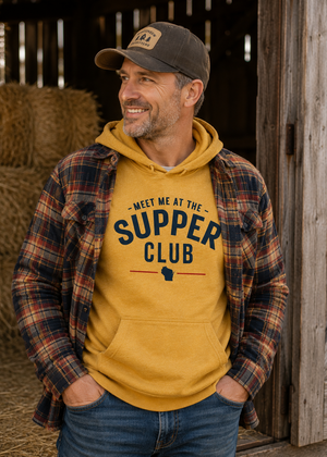 A man wearing a mustard yellow hoodie with 'MEET ME AT THE SUPPER CLUB' printed on the front, layered under an open red and navy plaid flannel shirt, blue jeans, and a dark baseball cap, standing in a rustic barn doorway.