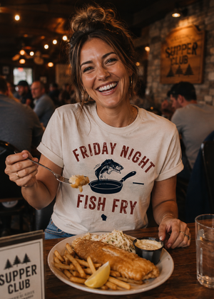 A woman smiling at a restaurant table while wearing a cream graphic t-shirt that reads 'FRIDAY NIGHT FISH FRY' with a fish in a pan illustration. In front of her is a plate of fish and fries, and the restaurant 'Supper Club' is visible in the background.