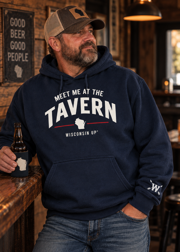 A man wearing a navy blue hoodie with 'MEET ME AT THE TAVERN WISCONSIN UP!' printed on the front, standing at a wooden bar in a rustic bar setting.