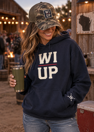 A woman wearing a navy blue hoodie with 'WI UP' printed on the front, a camouflage trucker hat with a 'WI UP' patch, and holding an olive green tumbler in an outdoor rustic setting.