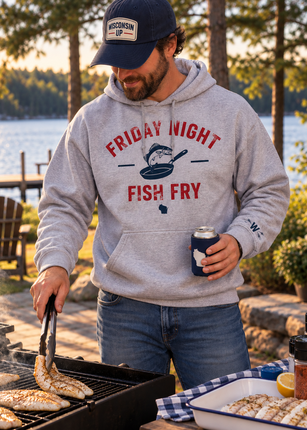 A man grilling outdoors by a lake wearing a light gray hoodie with a red and navy graphic that reads 'FRIDAY NIGHT FISH FRY'. He is also wearing a navy baseball cap with a 'Wisconsin Fish' patch.