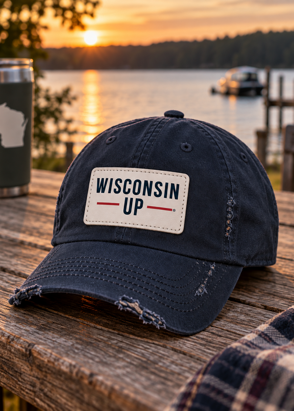 A distressed dark navy baseball cap with a white rectangular patch reading 'WISCONSIN UP' in navy and red, resting on a wooden table outdoors at sunset with a lake and dock in the background.