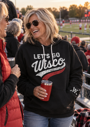 A woman at a football game wearing a black hoodie with 'LET'S GO Wisco' printed in white and red on the front, holding a red insulated tumbler.
