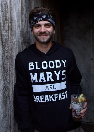 Man wearing a hoodie with 'Bloody Marys are Breakfast' text, holding a drink against a textured wall.