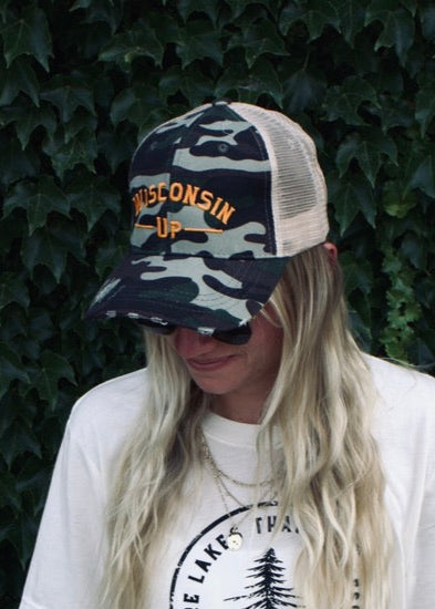 Person wearing a white t-shirt with a graphic design and a camouflage cap, standing against a dark green leafy background.