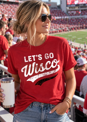 A woman in a stadium wearing a red graphic t-shirt that reads 'LET'S GO Wisco' in distressed white lettering with a black swoosh underneath. She is holding a white insulated tumbler and wearing sunglasses and denim jeans.