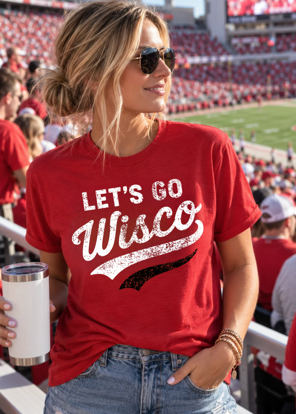 A woman in a stadium wearing a red graphic t-shirt that reads 'LET'S GO Wisco' in distressed white lettering with a black swoosh underneath. She is holding a white insulated tumbler and wearing sunglasses and denim jeans.