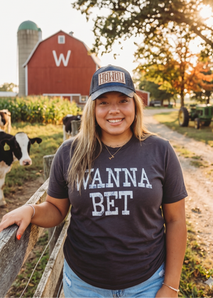 Woman wearing a 'Wanna Bet' shirt and cap standing in a farm setting with cows and a red barn.
