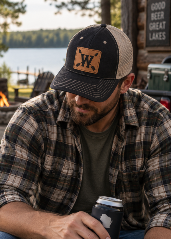 A man wearing a black and tan trucker hat with a leather patch logo sits outdoors by a lake, holding a can cooler.