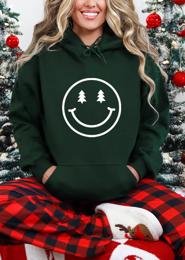 Person wearing a green hoodie with a smiley face design and red plaid pants in front of Christmas trees.