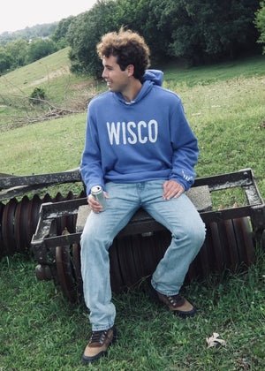 Person wearing a blue 'WISCO' sweatshirt sitting on a piece of farm equipment in a grassy field.