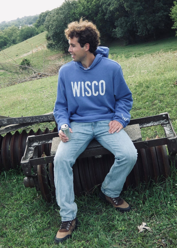 Person wearing a blue 'WISCO' sweatshirt sitting on a piece of farm equipment in a grassy field.