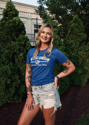 Woman wearing a blue 'Wisconsin Up' t-shirt standing outdoors with trees and a building in the background.