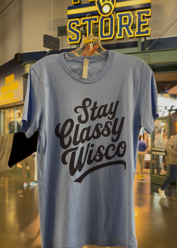 Light blue t-shirt with 'Stay Classy Wisco' text on a hanger in a store setting.