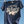 Load image into Gallery viewer, A navy blue graphic t-shirt hanging on a clothesline with wooden clothespins against a clear blue sky. The shirt features a large white graphic that reads 'IT MUST BE THE Wisco 2026 SERIES' with a baseball design.
