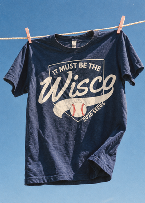 A navy blue graphic t-shirt hanging on a clothesline with wooden clothespins against a clear blue sky. The shirt features a large white graphic that reads 'IT MUST BE THE Wisco 2026 SERIES' with a baseball design.